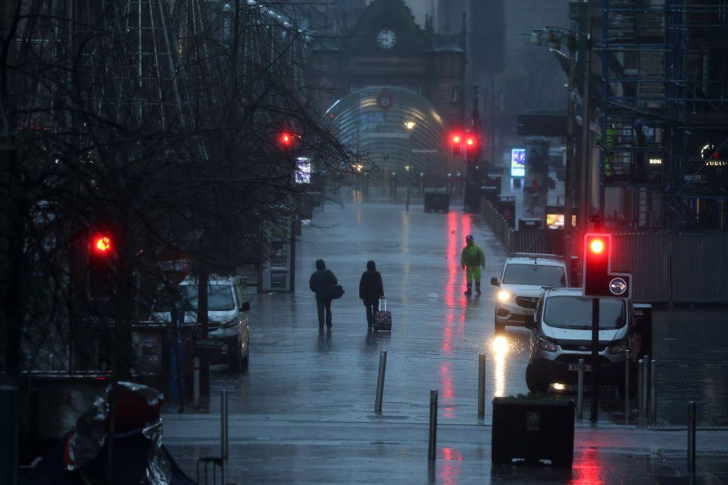 Buchanan Street in Glasgow, looking down towards the entrance to St Enoch's underground station. It is dayight though very dark. There are red traffic lightts reflecting in the wet street and two people can be seen walking along with bags at a distance. 