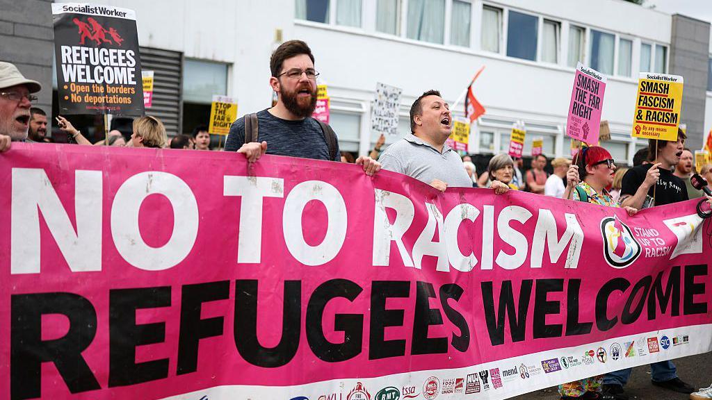 A counter-protest outside the Cladhan Hotel in Falkirk on 16 August. A row of people are holding a pink banner which reads No To Racism and Refugees Welcome, and carrying placards.