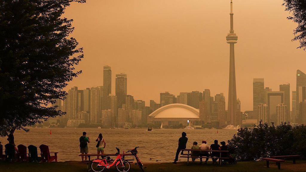 An image showing the Toronto skyline taken from the Toronto Islands showing a hazy, orange sky blanket the city. On the shores of the island are several people sitting on benches, and a couple of city bikes parked nearby. The skyline across the lake shows the iconic CN Tower and Rogers Centre.
