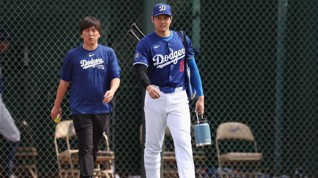 Ippei Mizuhara, the former interpreter to Los Angeles Dodgers's Shohei Ohtani, seen walking with the baseball star on the field. He is wearing a blue Dodgers shirt and Shohei is in his baseball uniform. 