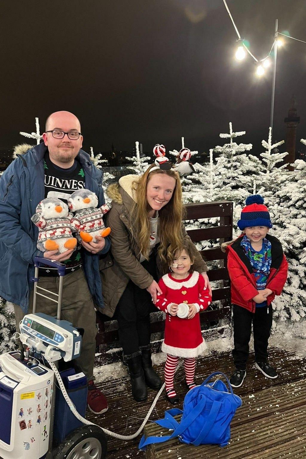 The Green family at a Christmas event in 2023. Evie is wearing a red Christmas outfit and is attached to a Berlin Heart machine. Her brother Theo is to her left, and their parents, David and Chloe, are next to them. Mr Green is holding two toy penguins. White Christmas trees are in the background of the photo.