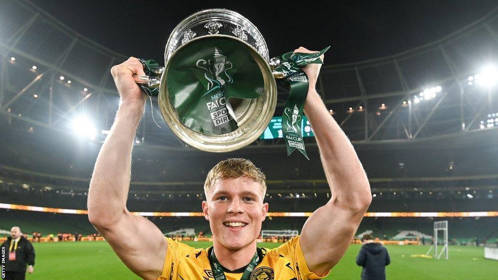 A man in a yellow football shirt holds a silver trophy above his head. The trophy has green ribbons on it and the player is wearing a medal tied with similar green ribbon around his neck. He has short blonde hair and is smiling. He is on the pitch of a large floodlit stadium.