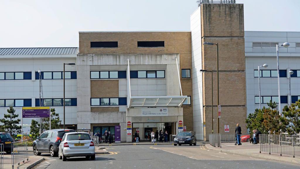 The Royal Infirmary of Edinburgh hospital. It is a white and brown brick building. Cars are driving towards the entrance and there are people walking along the pavements.