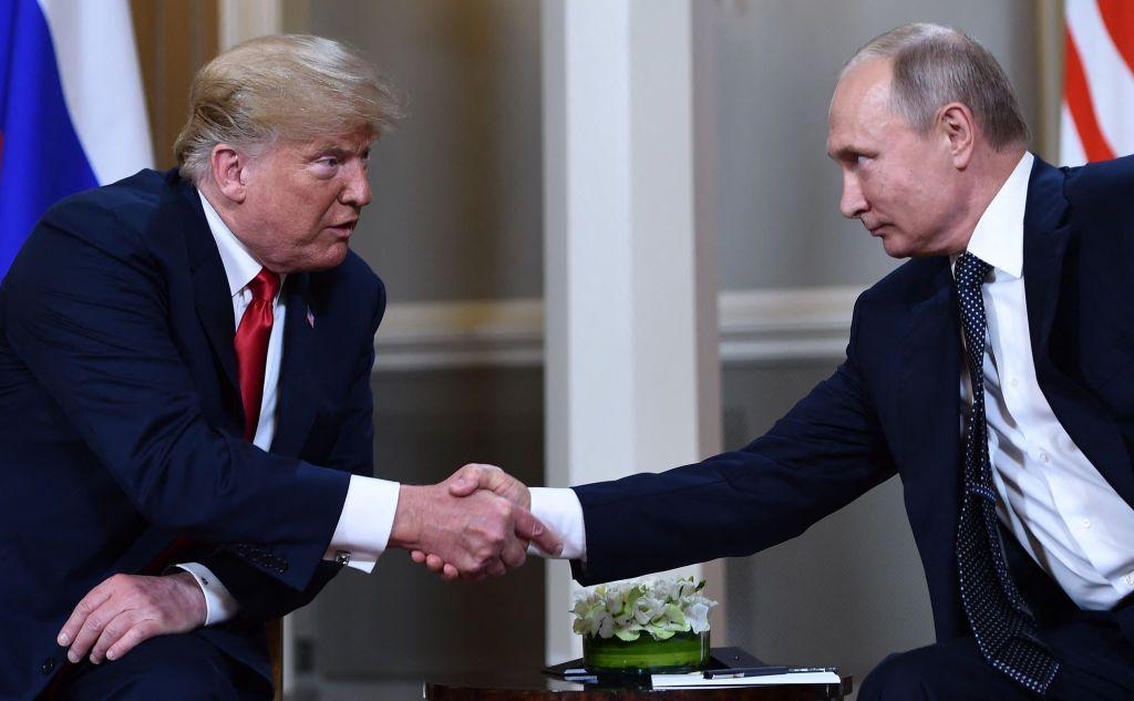 Russian President Vladimir Putin and US President Donald Trump shake hands before a meeting in Helsinki. They are both sitting down, wearing dark suits and Donald Trump is saying something to Putin.