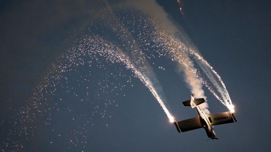 A plane flying in the night sky upside-down. It has pyrotechnics coming from the tips of its wings