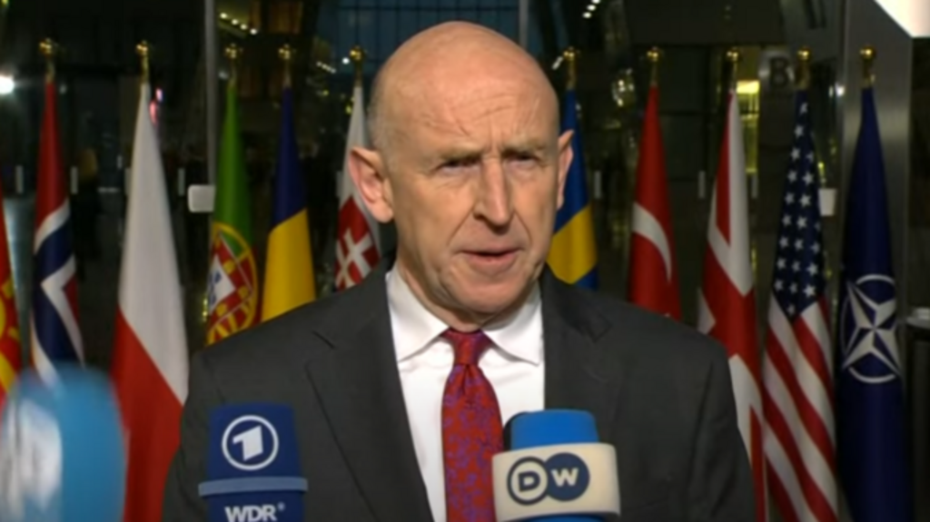 Defence Secretary John Healy answers questions ahead of Nato summit, two mics in front of him as he stands in dark grey suit, white shirt and red tie