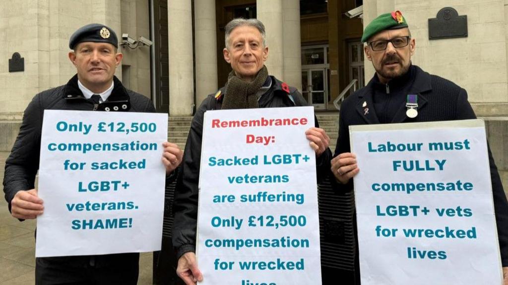 Three men, two of whom are armed forces veterans standing in a row holding placards that have messages about £12500 compensation for LGBT+ veterans not being enough. The veteran on the right is Adrian Radford-Shute.