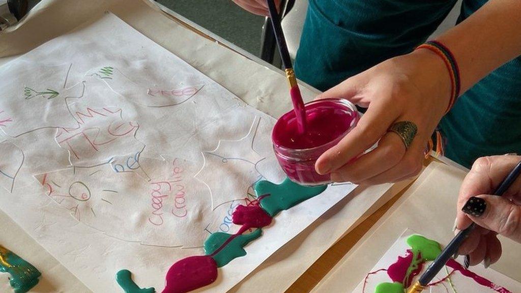 A piece of artwork with a person's hands in shot, dipping a paintbrush into pink paint