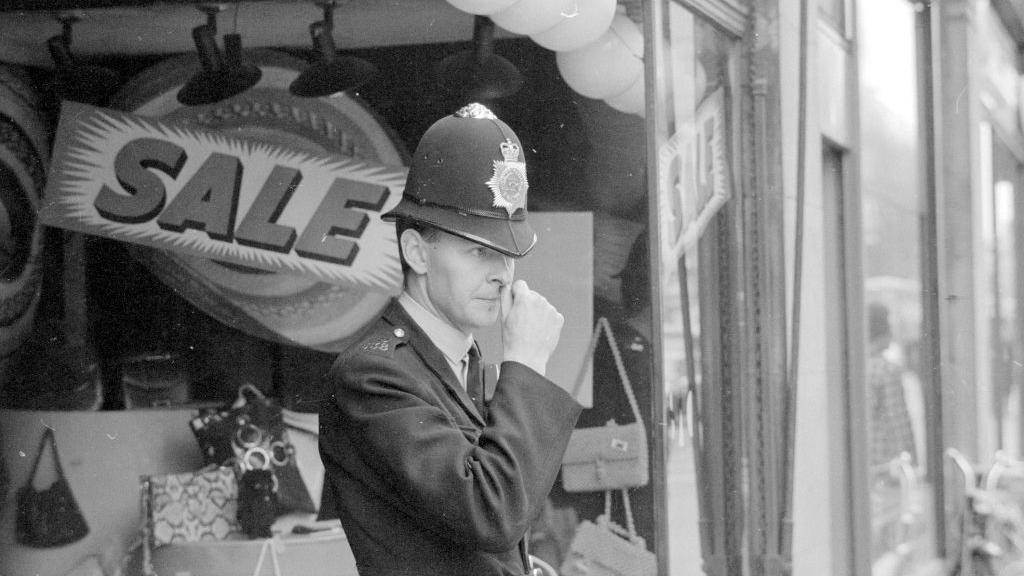 A uniformed police officer stands outside the SAC shop. He is wearing a tall helmet and is scratching his nose with his thumb