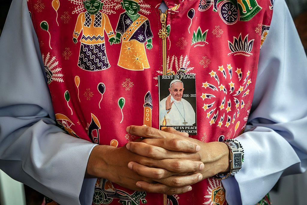 A Catholic nun prays during a Requiem Mass in honour of Pope Francis. She wears a red, patterned tunic and pinned to her clothing is a portrait of the late Pope.