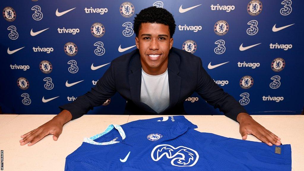 New Chelsea signing Andrey Santos smiles as he sits at a desk with a blue Chelsea shirt on it