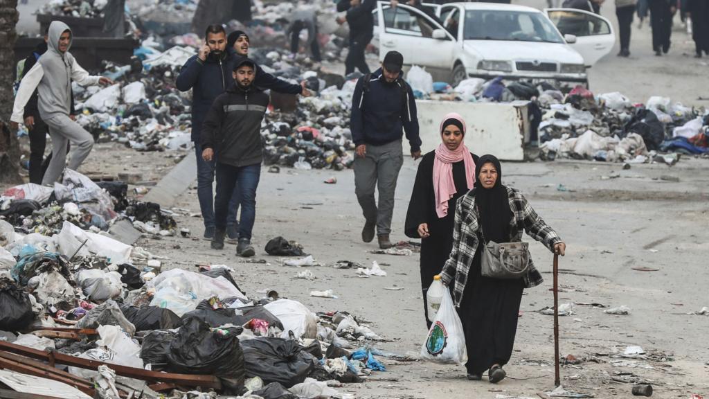 Palestinians walk among rubble in Gaza City - a woman with a walking stick, holding a plastic bag with some belongings in it, leads the way.