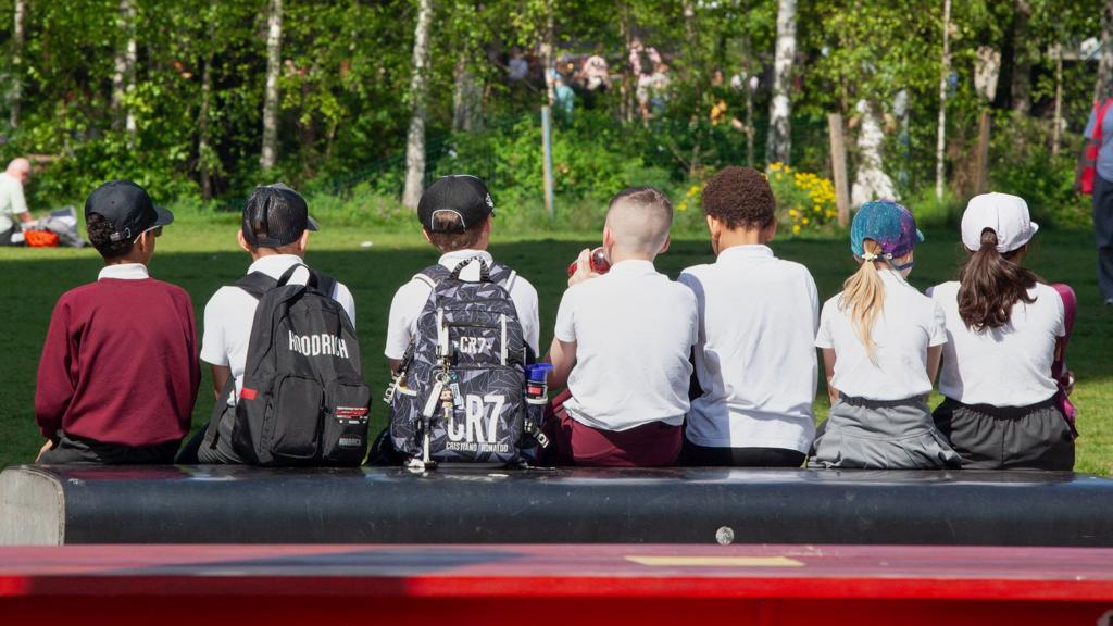 Schoolchildren sit in the sun while wearing protective sun hats and wearing backpacks and school uniform, with their backs to the camera so they are not identifiable, sitting on a bench outside while on a school trip to the Tate Modern on 1 May