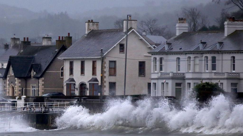 Waves break against a sea wall. Behind the wall and road and on the other side of the road houses that are grey, white and brown.