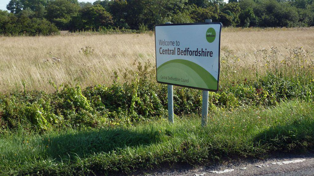 A sign reading: "Welcome to Central Bedfordshire", by the side of a road, with grass behind it in a field, a bush and more green grass. 