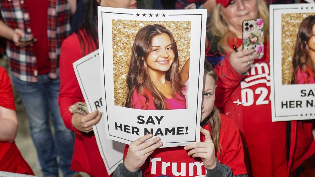 Women wearing red Trump 2024 T-shirts hold up signs with Laken Riley's photo and the words "say her name!"