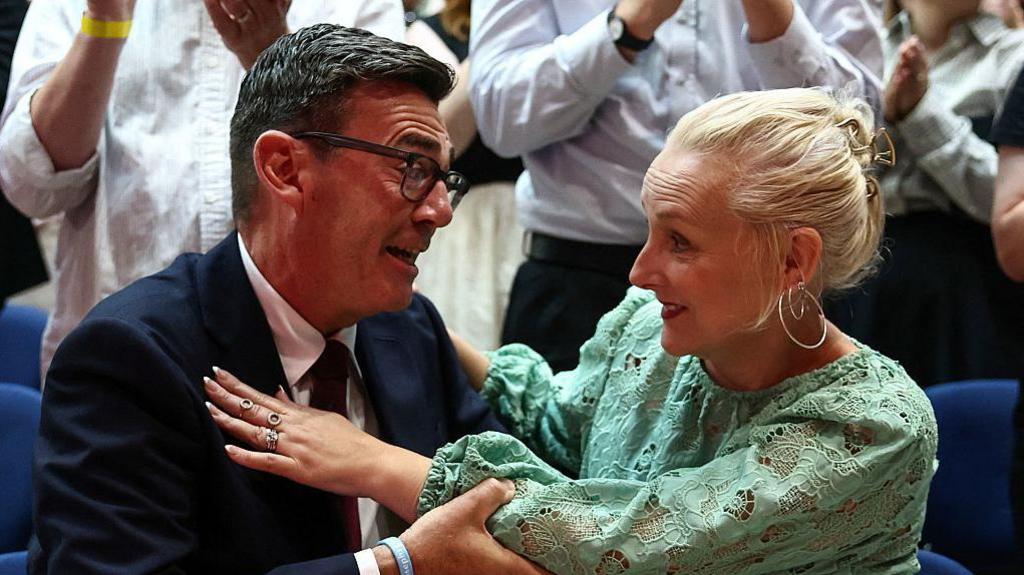 Andy Burnham hugging his wife, Marie-France van Heel. Burnham is wearing a dark suit with a shirt and tie and glasses. Van Heel is wearing a green dress. She has blond hair and hooped earrings. 