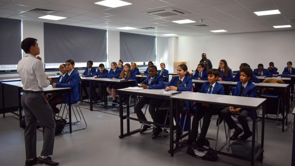 A classroom with pupils sat at desks facing the camera, and a male teacher stood at the front with his back to the camera wearing a white shirt and grey trousers