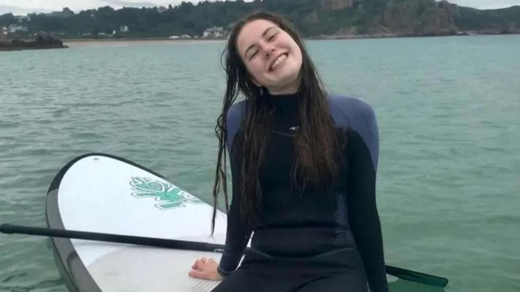 Lily sat on a paddle board in a wetsuit smiling at the camera. Her long dark hair is wet from the sea. 