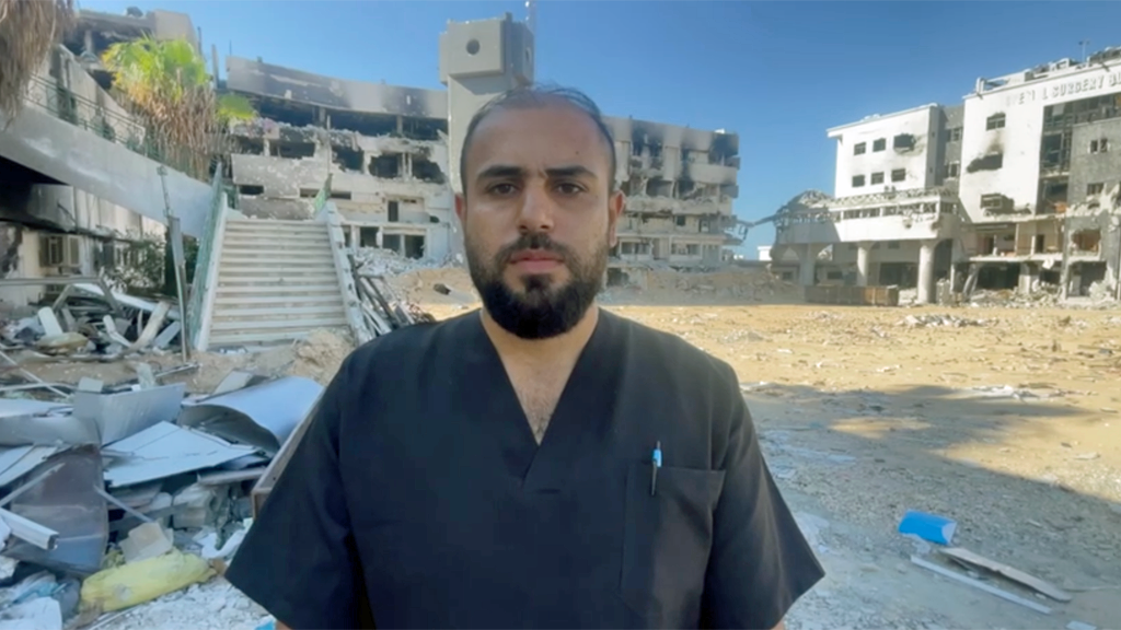 Dr Elawa, a man with thinning dark hair and a beard, stands looking into the camera, wearing black v-neck hospital scrubs with a pen in the pocket. In the background are buildings damaged by explosions, with scorch marks and debris all around the ground. The sky is blue.