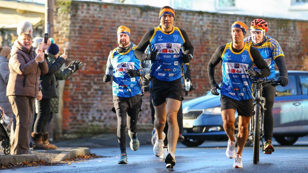 Kevin Sinfield runs through a street flanked by a support crew and cheered on by members of the public.
