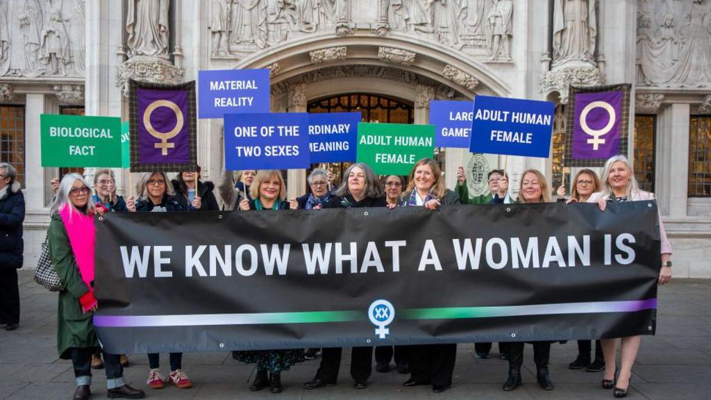 For Women Scotland campaigners outside the Supreme Court, holding up a banner reading "we know what a woman is". There are 14 women standing in a row, many holding up smaller banners.