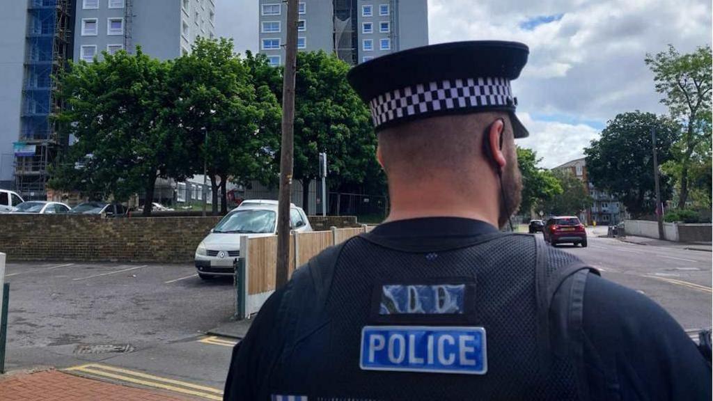 Upper body and head of a police officer with his back to the camera. He is wearing a black shirt with POLICE in white letters on a blue background. He is also wearing a black cap with white and black braiding. He has very short dark hair and a beard. He is looking at a wide road on which a red car is visible. To his left are a tall building, trees, a stone fence and parked cars.