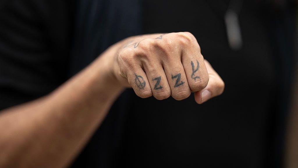 A close-up photo of a hand in a fist shape, with the word "Ozzy" tattooed across the four fingers. There is one letter on each of the person's fingers.  