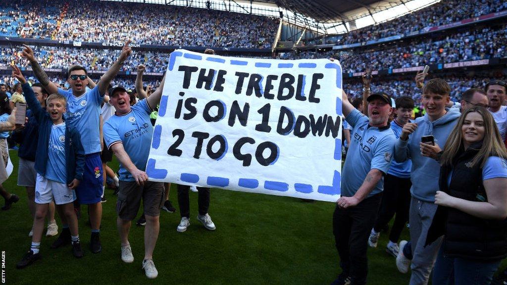 Manchester City fans on the pitch display a banner with "The Treble is on, one down two to go" written on it