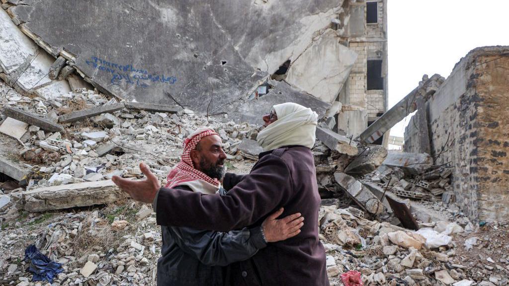 Two men in scarves embrace in front of a building turned into rubble in Baba Amr district of Homs on 6 December