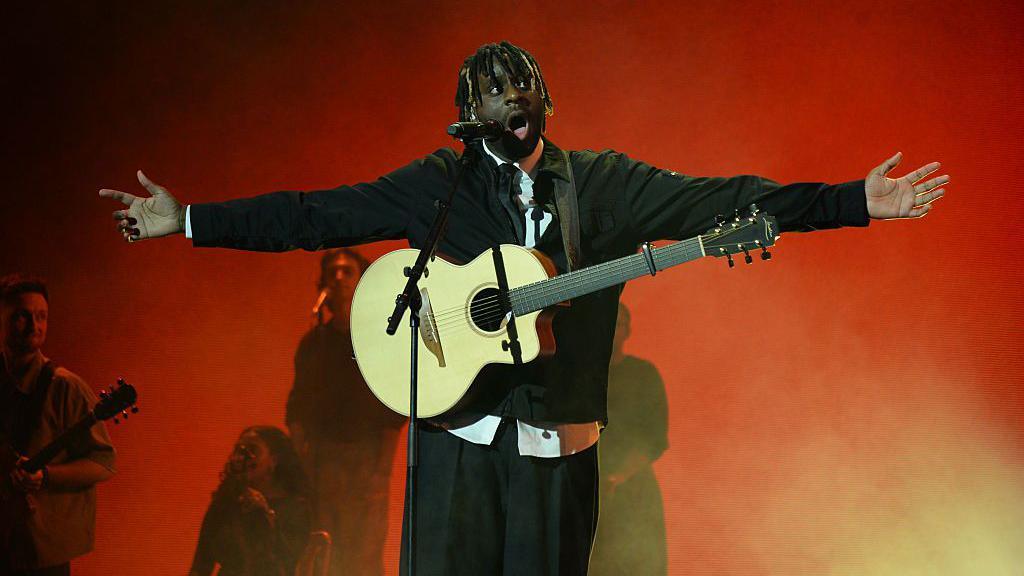 Myles Smith standing on stage with his arms spread wide, an acoustic guitar hanging round his neck