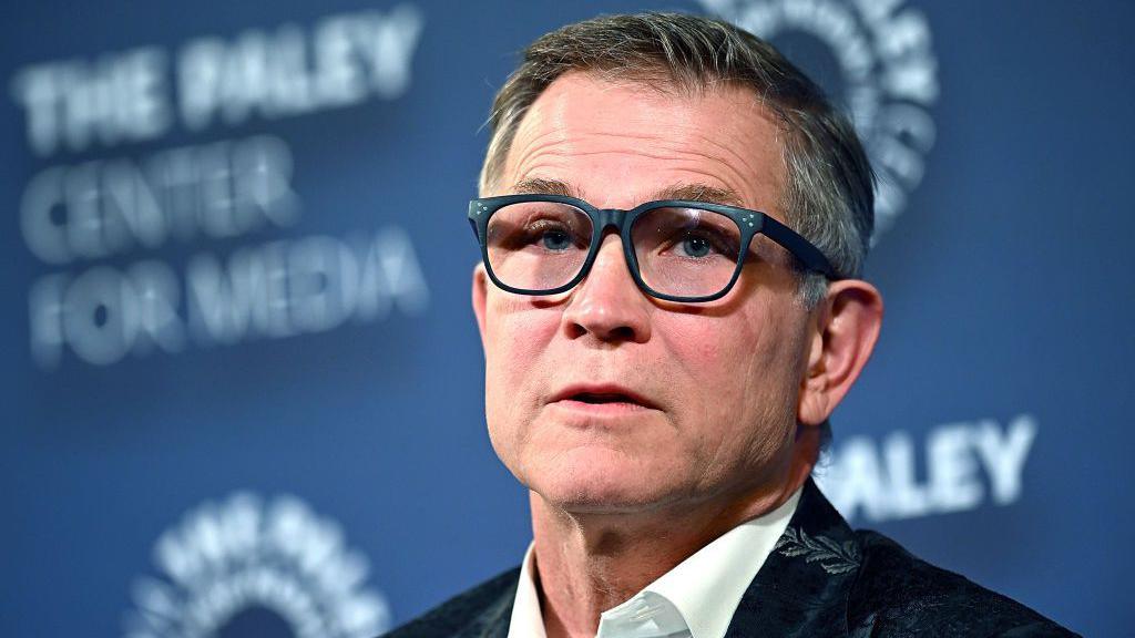 Man with grey and black hair in glasses and in a suit with leaf patterns has his mouth slightly open as he looks towards the camera