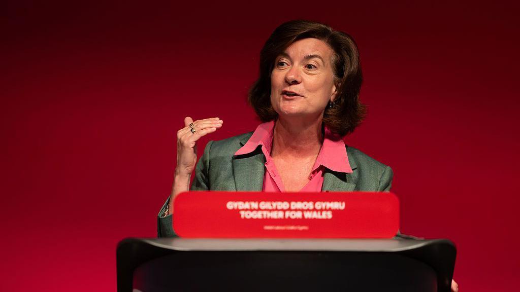 Eluned at the podium with a labour slogan in white print on read that says Together for wales and the Welsh language translation. There is also a red background behind the stage. Eluned has brown shoulder length hair and is wearing a pink open neck blouse with a green business jacket. She is gesturing with on hand as she enunciates a word that has her mouth closed and her brow slightly raised. 