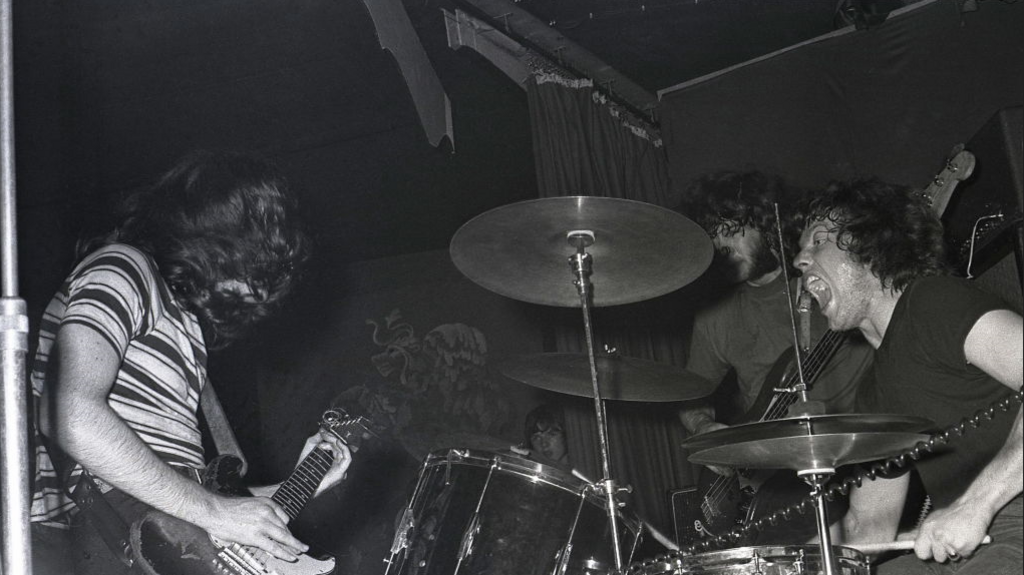 Rory Gallagher in striped t-shirt, with John Wilson on drums, and Richard McCracken, performing at a gig in Germany