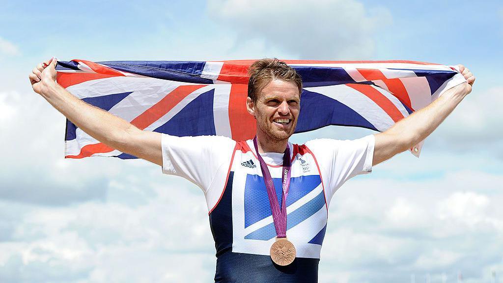 Alan Campbell holding the union flag above his head, wearing a bonze medal