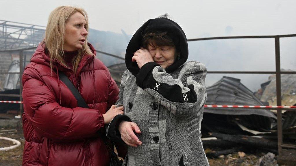 Two women stand in front of a damaged building