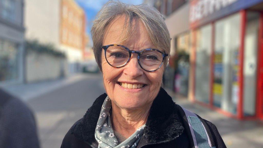 A head-and-shoulders photo of Jane Morgan, a woman with short grey hair and glasses and wearing a black coat, smiling at the camera with a parade of shops behind her.