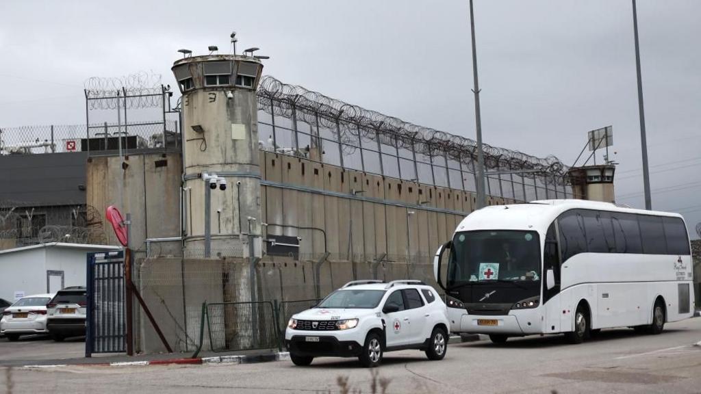 A bus and car wait outside a prison with high walls and railings.