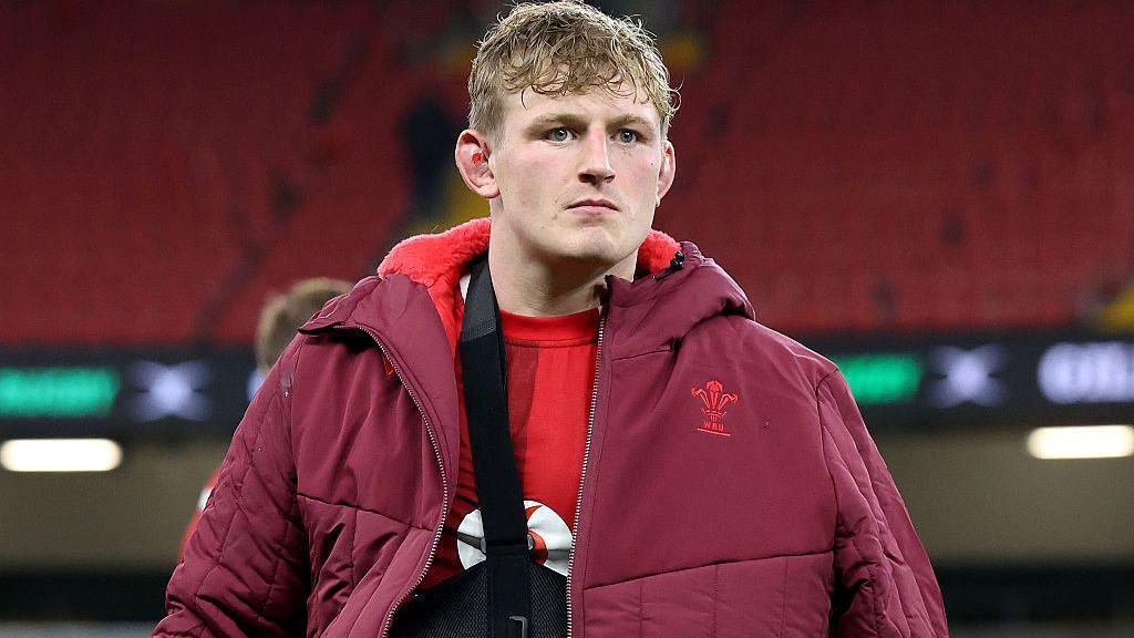 Jac Morgan wearing his arm in a sling under a red Wales-branded coat