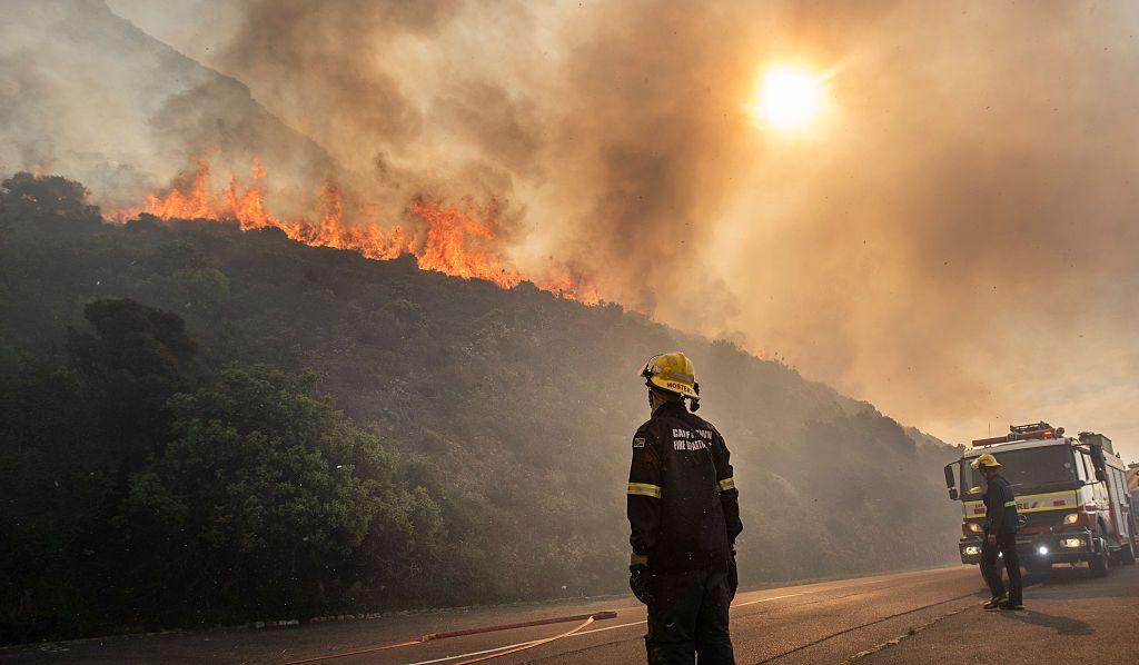 Firefighters look on as a fire blazes above the roadside. They have lorries and hose pipes which they are readying for use.