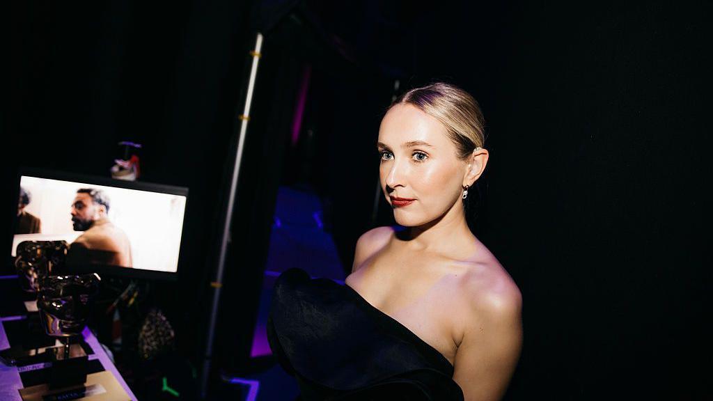 A glamorous woman in a black dress stands in the darkness behind a stage.