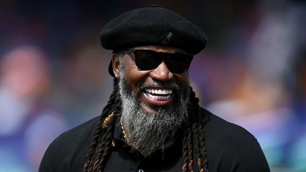 Former West Indies cricketer Chris Gayle wearing sunglasses and a black jersey and widely smiling.