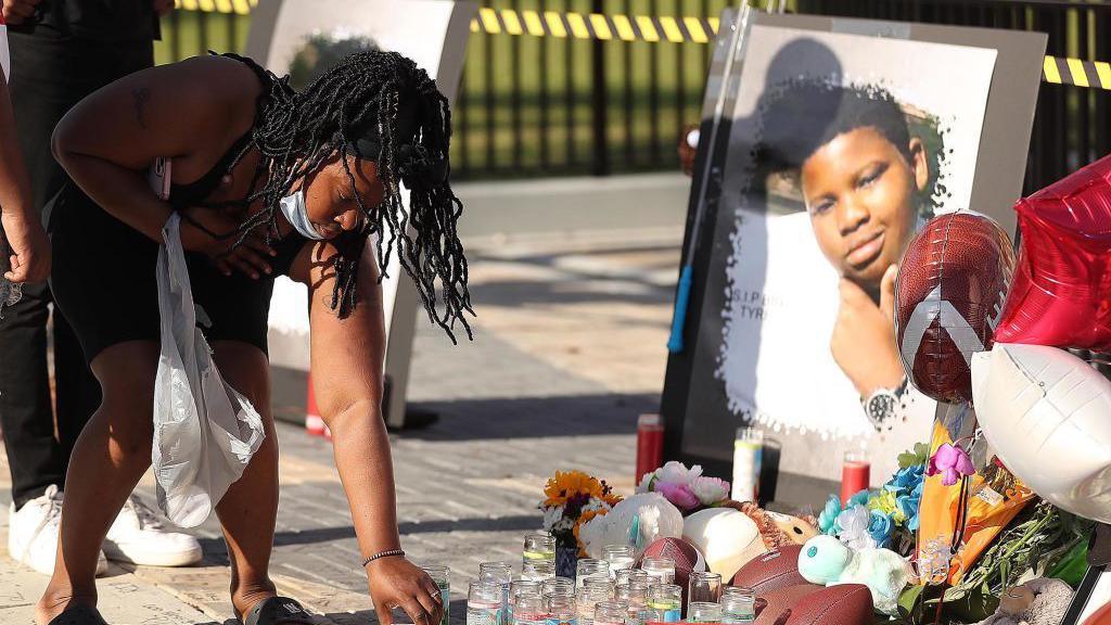 A person placing a candle beside flowers, balloons and a picture of Tyre 