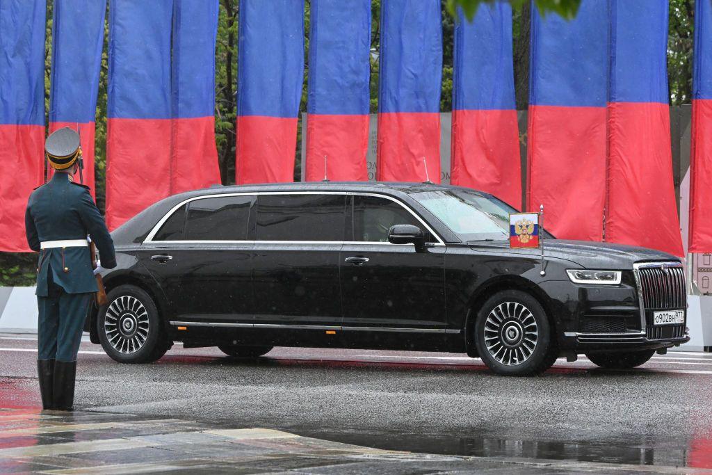 An Aurus Senat car drives in front of flags and behind a figure in military uniform