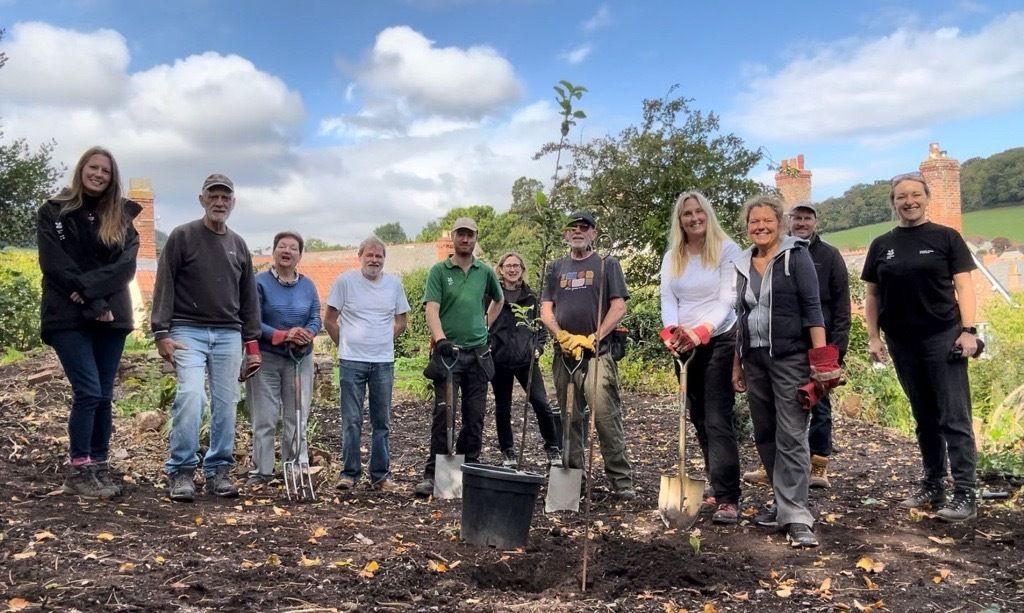 A group of 12 people are standing in a muddy field, with many of them holding shovels and forks. They are all looking at the camera, smiling.