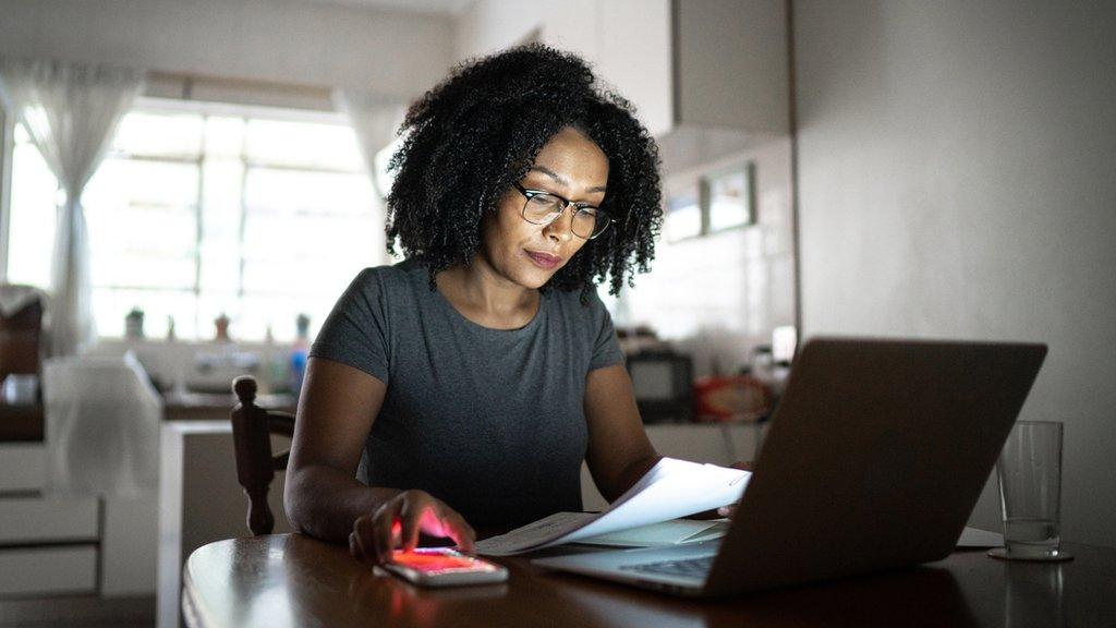 Woman looks at bills while holding a smartphone in front of a laptop