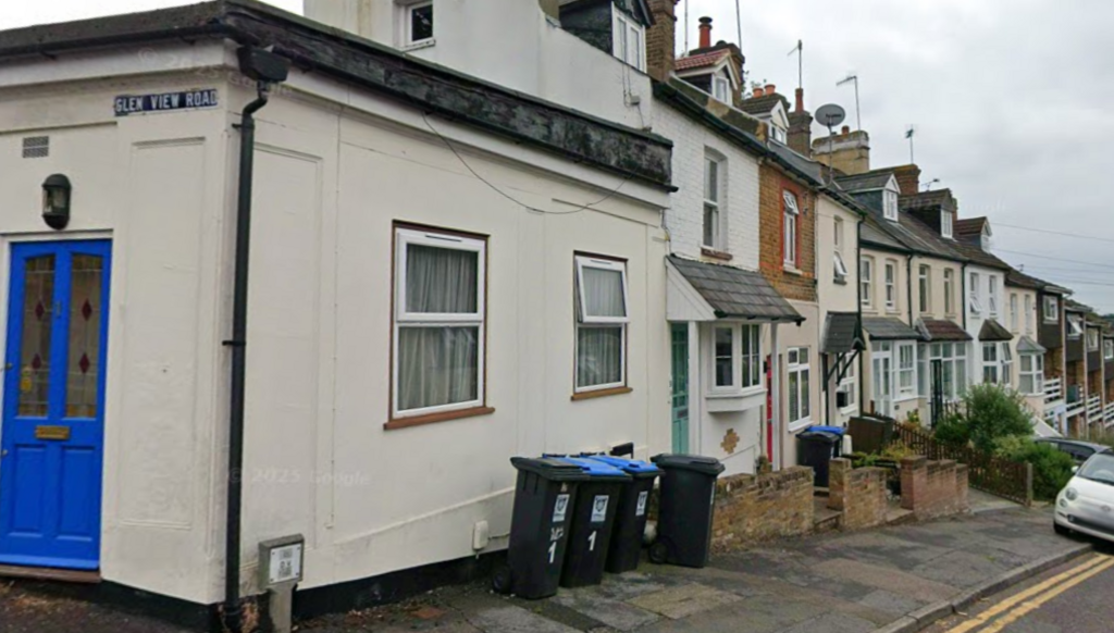A row of houses on a street which is on a slight hill. There are bins outside the houses and there is a white car parked half on the pavement. There is a sign on the corner saying "Glen View Road"