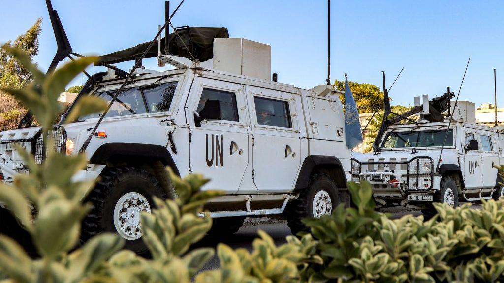 Two white Armoured UNIFIL vehicles, with some green bushes in the foreground and blue skies in the background. 