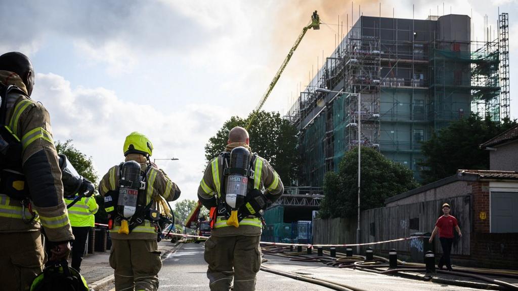 Firefighters approach the burning building, with other firefighters dousing the blaze from a crane ladder