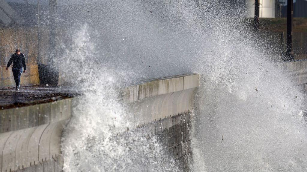 A man walks along the shore path in Saltcoats as a huge wave rises up above the harbour wall and almost submerges him